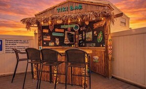 Eine Tiki-Bar mit einem Reetdach, Holzfassade und einem Schild, das sie als Fintik Bar kennzeichnet. Vor der Bar stehen drei schwarze Metallstühle.