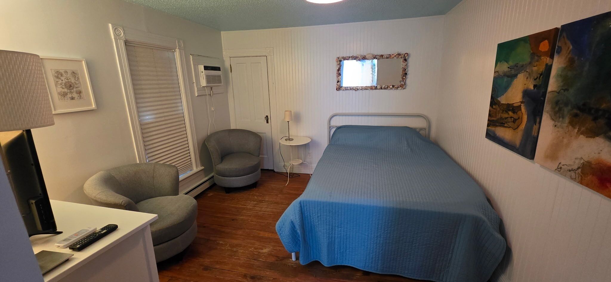 A bedroom with a bed, two armchairs, a desk with a lamp, a small table, and a painting on the wall.