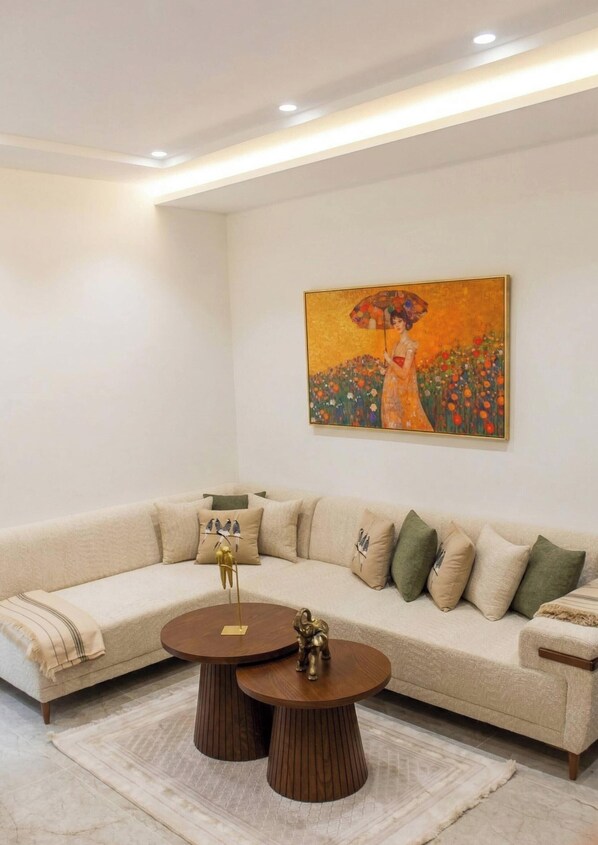 Una sala de estar moderna con un sofá seccional color beige, dos mesas redondas de madera y un cuadro grande de una mujer sosteniendo un paraguas.
