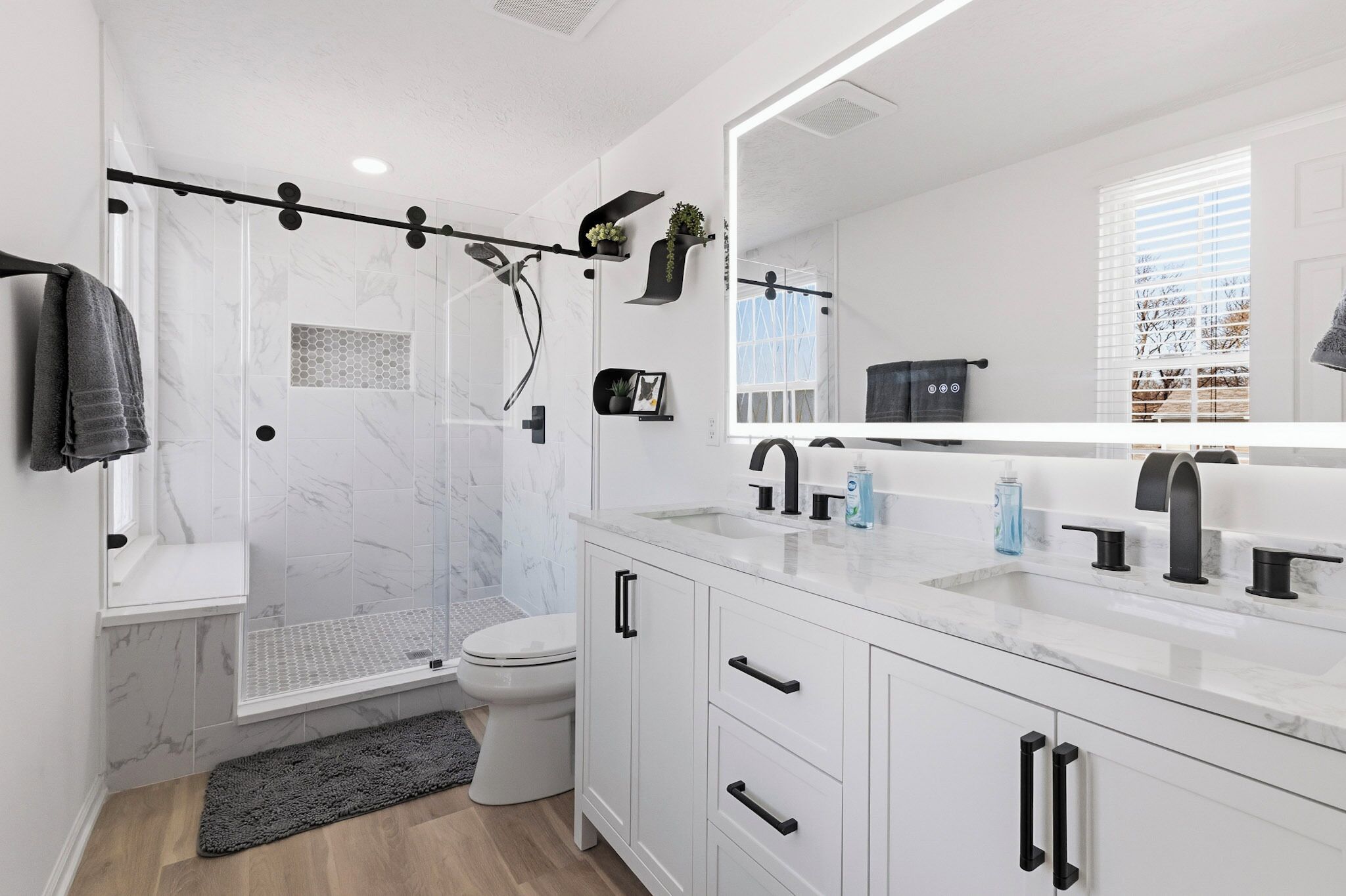 A modern bathroom with a glass shower enclosure, white marble tiles, a large mirror, and a white vanity with black handles.