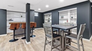A modern dining area with a wooden table and chairs, a bar area with stools, and built-in shelves with decorative items. - Luxury Fishers,IN Home - 4BR • Fully Renovated • Finished Basement • WiFi (Fishers)