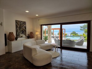 A modern living room with a sectional sofa, a dining area, and a view of an outdoor pool and garden. - Villa Muse Sea View and private Pool (Afitos)