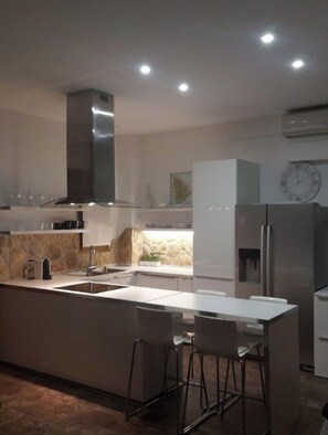 A modern kitchen with a central island, bar stools, a built-in oven, and a clock on the wall. - Villa Muse Sea View and private Pool (Afitos)