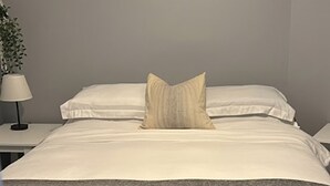 Un letto rifatto con lenzuola bianche e un solo cuscino beige, una lampada da comodino e una pianta sul lato sinistro.