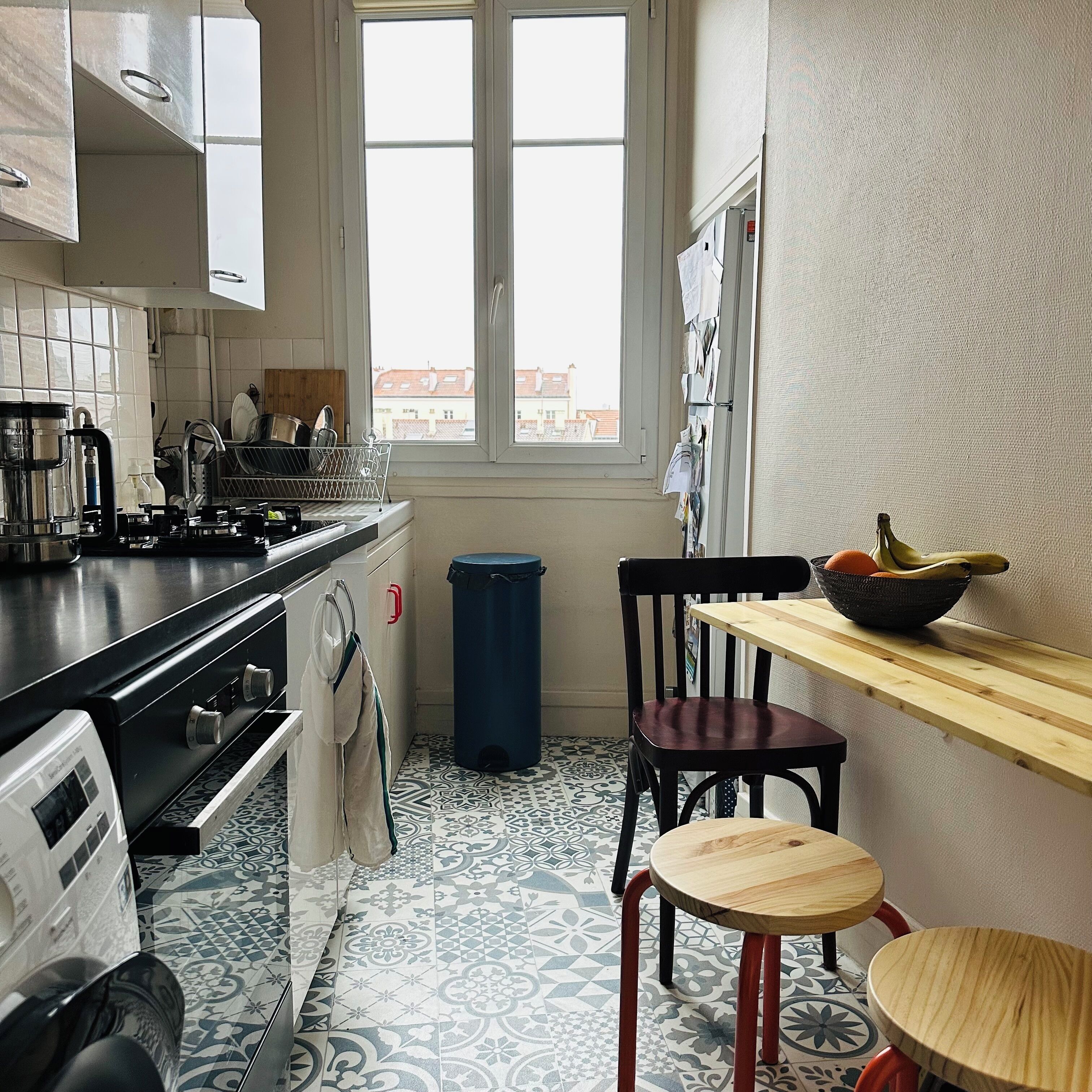A kitchen with a stove, sink, and window overlooking buildings. There is a trash bin and a chair near the window.