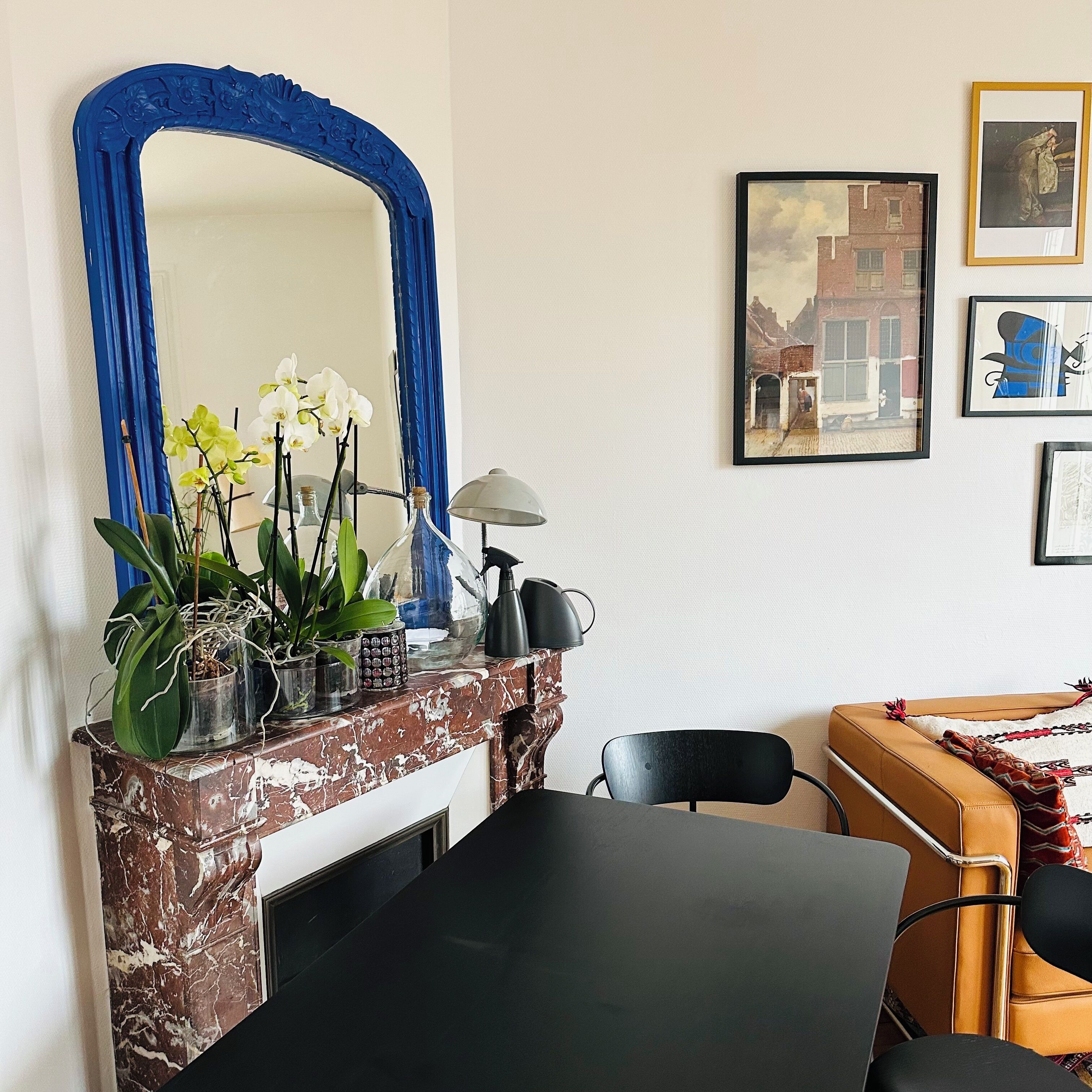A room with a marble fireplace, a blue-framed mirror, a table, chairs, and artwork on the wall.