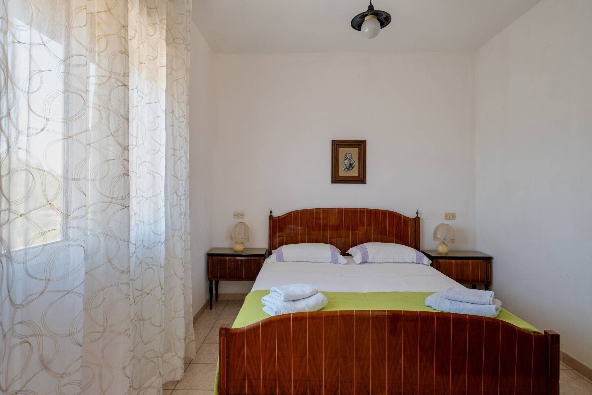 Une chambre à coucher avec un lit en bois, deux tables de chevet avec des lampes, un tableau encadré au mur et une fenêtre avec des rideaux légers.