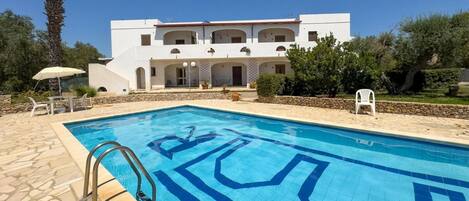 A swimming pool with a geometric tile pattern, a white building with multiple balconies, and a stone pathway leading to the pool.
