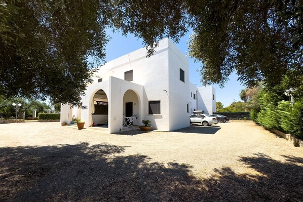 A white building with arched entrances, a gravel driveway, and parked cars.