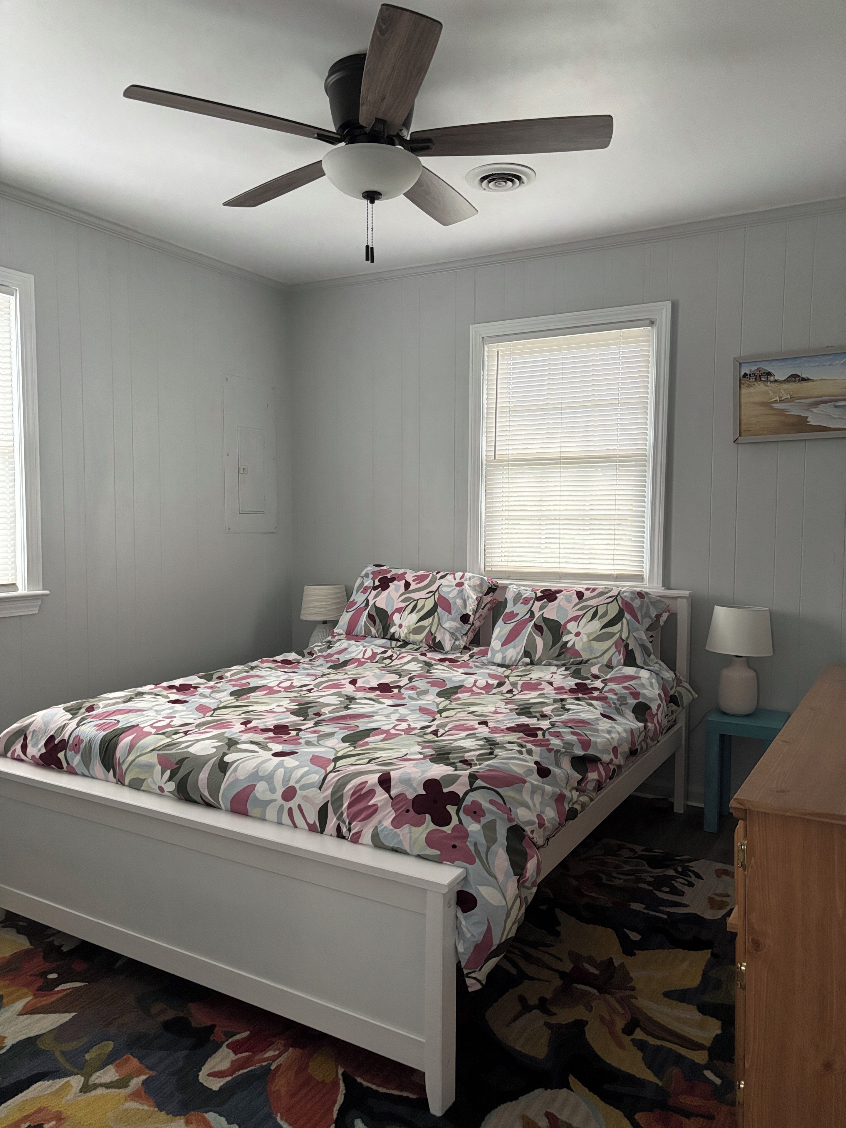 A bedroom with a bed, a ceiling fan, a window with blinds, a bedside table, and a lamp.