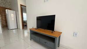 Una habitación con televisión sobre un mueble de madera, un refrigerador y piso de baldosas.