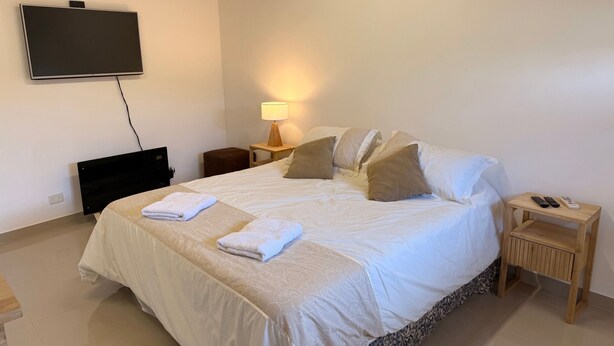 A neatly made bed with white linens and two pillows, a bedside table with a lamp, a TV mounted on the wall, and a small wooden stool with a remote control. - Ezeiza Victoria Barrio Uno, only 5' min from Ezeiza airport. (Ezeiza)