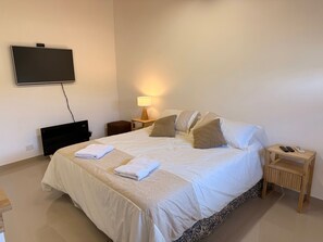 Uma cama bem arrumada com roupa de cama branca e duas almofadas, uma mesa de cabeceira com abajur, uma TV fixada na parede e um banquinho de madeira com controle remoto.