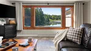 A living room with a large window, a leather sofa, a coffee table with books, and a flat-screen TV. - Private Lakefront House, 15 feet to water, Kayaks, Game Room, Dog Friendly (Wolverine)