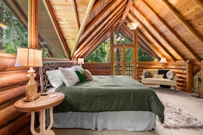 Ein Schlafzimmer in einer Blockhütte mit einer hohen Decke, einem Bett mit grüner Bettwäsche, einem runden Holztisch und einem Sofa.