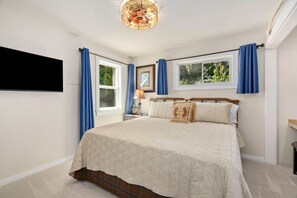 A bedroom with a bed, a nightstand, a lamp, a wall-mounted TV, and two windows with curtains. - Amazing Seattle view beachfront 4 bedroom /3 bath (Bainbridge Island)