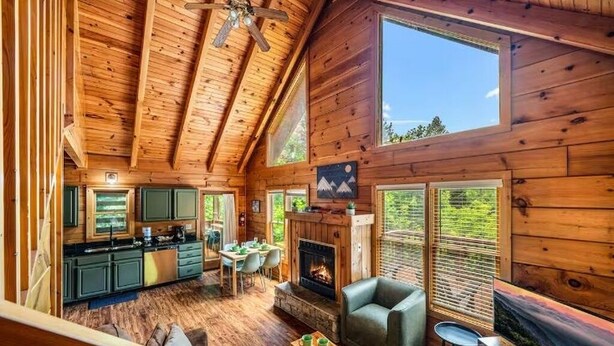 Interior de una cabaña con comedor, cocina, chimenea y ventanas amplias.