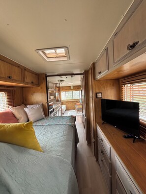 Interior compacto de una caravana con cama, kitchenette y televisor.