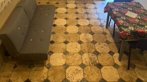 Een knus vertrek met een stenen plafond, een bank, een eettafel met een bloemrijk tafelkleed, zeshoekig patroon vloertegels en een kast met decoratieve objecten.
