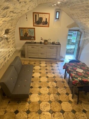 Ein gemütliches Zimmer mit Steindecke, einer Couch, einem Esstisch mit Blumenmuster-Tischdecke, sechseckigem Fliesenboden und einer Vitrine mit Dekoration.