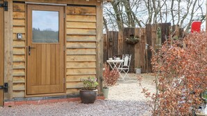 A wooden cabin with a door, a small patio with a table and chairs, and a gravel ground. - Charming apartment set in the Wye Valley, Herefordshire (Perrystone Hill, Ross-on-Wye)