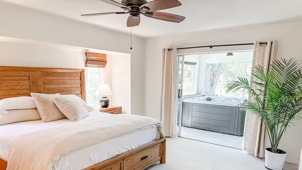 Une chambre avec un lit en bois, un ventilateur de plafond, une fenêtre avec des stores, une table de chevet avec une lampe et une vue sur un jacuzzi, visible à travers une porte coulissante.