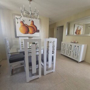 A dining area with a white table, chairs, and a chandelier. There is a painting of fruit on the wall and a cabinet with a mirror. - Las Palmas Residential Complex (Santiago de los Caballeros)