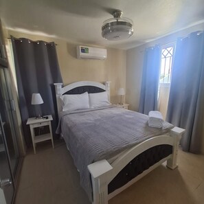 Ein ordentlich eingerichtetes Schlafzimmer mit Bett, Nachttischen, einer Lampe, einer Klimaanlage und einem Fenster mit Vorhängen.