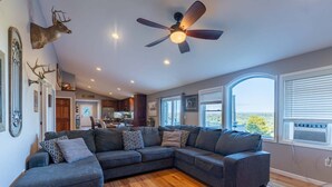 Un salon moderne avec un canapé d’angle, un ventilateur de plafond et de grandes fenêtres donnant sur un beau paysage.