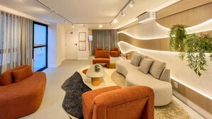 A modern living room with a sofa, armchairs, and a coffee table. There is a large window with curtains, a door, and a wall-mounted plant. - Saint Bartz Residence - Ondina (Salvador)