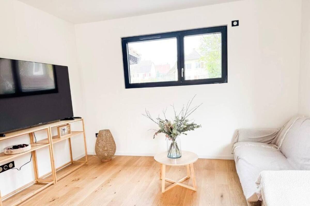 A living room with a TV on a wooden stand, a round wooden table with a vase, a sofa, and a window with blinds.