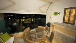 Ein Balkon mit Glastisch und Korbstühlen, eine Fischdekoration an der Wand und Blick auf die beleuchtete Stadt bei Nacht.