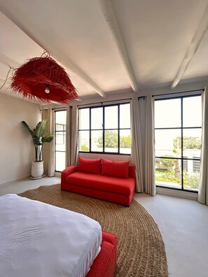 Ein modernes Schlafzimmer mit einem großen Bett, einem roten Sofa, einer Topfpflanze und Blick durch große Fenster.