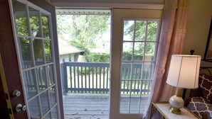 Une chambre avec vue sur une terrasse en bois, une porte blanche, une lampe sur une table de chevet et un coussin à motifs.