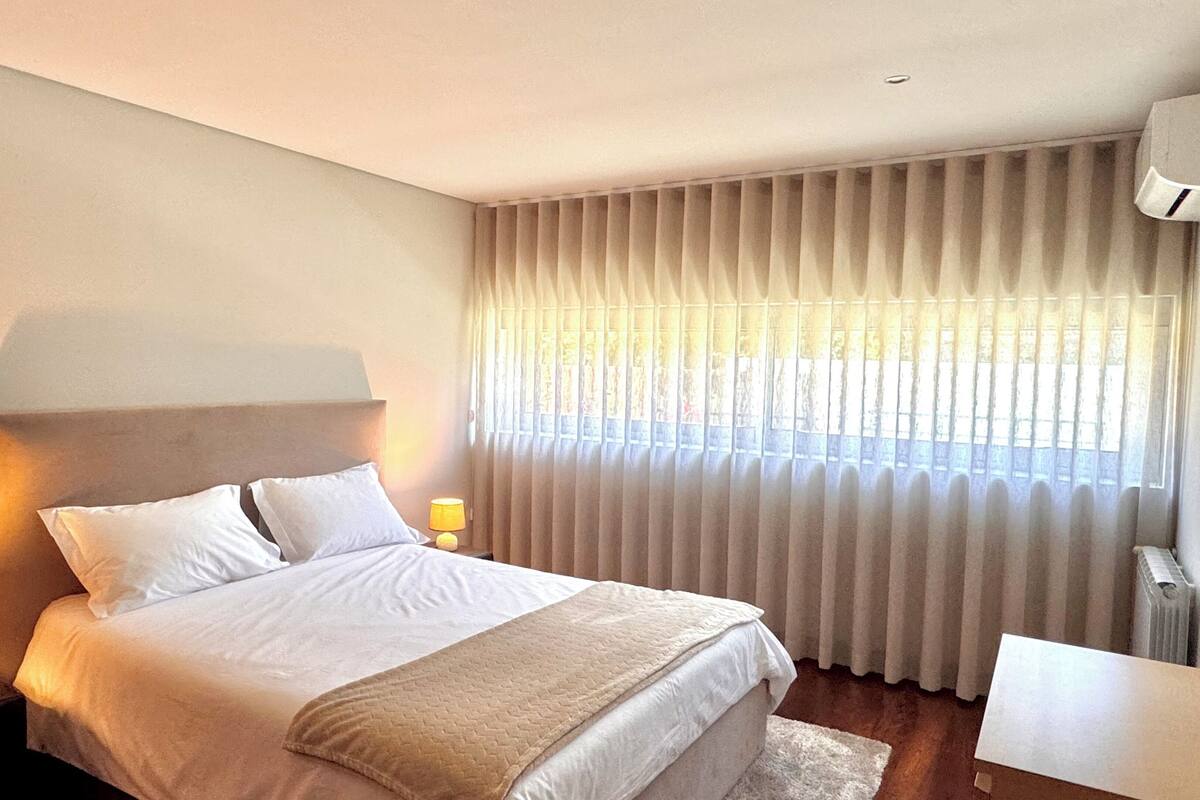 A bedroom with a bed, two bedside lamps, a window with curtains, a radiator, and a small table.