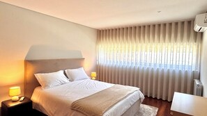 A bedroom with a bed, two bedside lamps, a window with curtains, a radiator, and a small table.