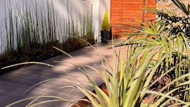 Un bâtiment moderne doté d’une porte en bois, entouré d’un jardin où poussent différentes espèces de plantes et bordé d’un sentier.