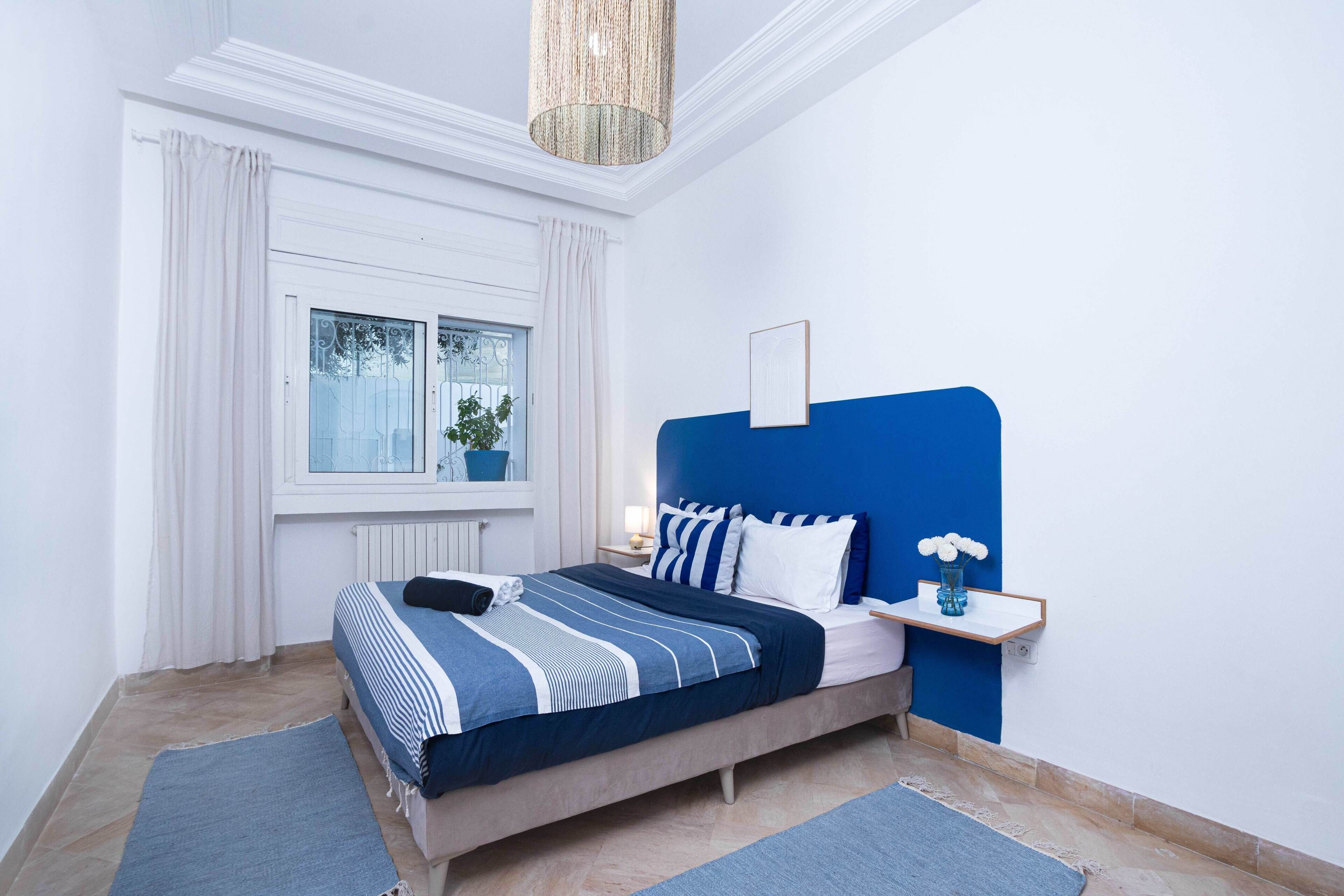 A bedroom with a bed, a nightstand, a window with curtains, and a hanging light fixture.