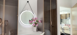 A bathroom with a round mirror, a vase of flowers, and a shower area. - Delightful 2-bedroom apartment with AC in Lapu-Lapu (Lapu-Lapu)