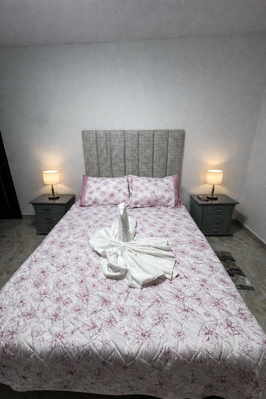 En snyggt bäddad säng med ett blommigt överkast, två sänglampor och en sänggavel.