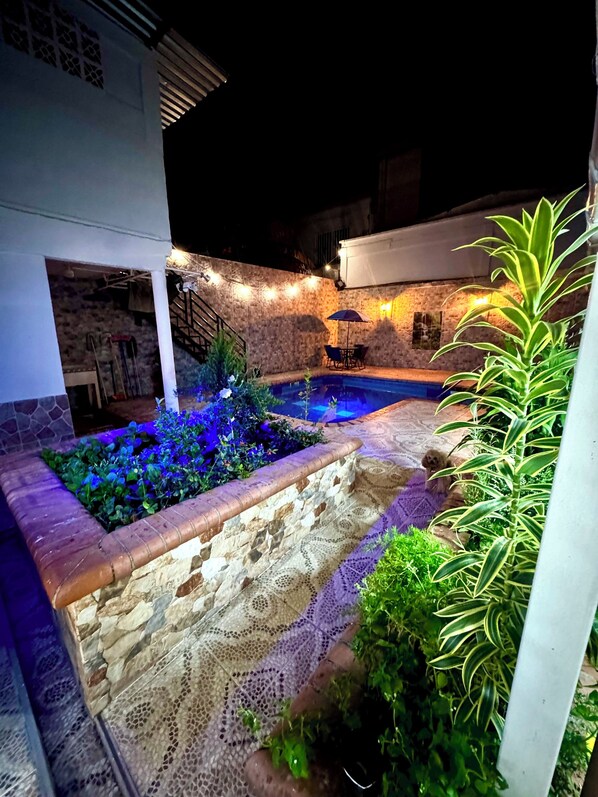 Un espacio exterior nocturno con piscina, paredes de piedra e iluminación de guirnaldas.
