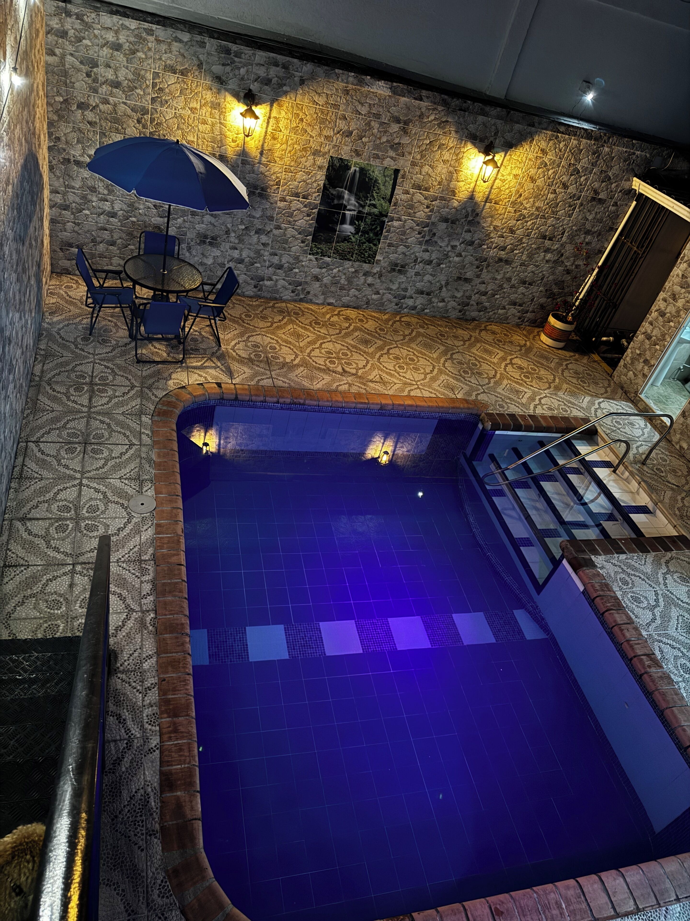 Una piscina en la azotea con suelo de baldosas azules, rodeada de baldosas estampadas y una pared de piedra. Hay una mesa con sillas y un paraguas azul.