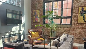 A living room with a brick wall, a large window, a sofa, a TV, and a potted plant. - Cozy 1-bedroom condo in Atlanta walking distance to Mercedes Benz Stadium (Atlanta)
