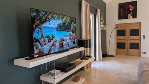 A modern living room with a flat-screen TV mounted on a white console, a wooden floor, a white sofa, a standing lamp, and a door with a glass panel. - Wine estate | 20m pool and cinema (Pézenas)