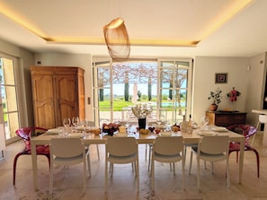 Comedor con una mesa dispuesta para seis personas, sillas, lámpara colgante y vista a un jardín a través de amplias ventanas.