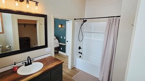 Ett badrum med ett handfat i trä, en vit tvättställ, en stor spegel, en dusch med duschdraperi och en hall som leder till ett sovrum.
