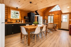 Ein Holzblockhaus-Interieur mit einem Holzesstisch, Stühlen, einer Küchenzeile inklusive Mikrowelle und einem großen Fenster.