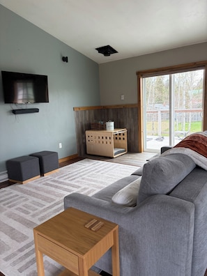 Ein Wohnzimmer mit grauem Sofa, einem Holztisch, einem flachbildfernseher an der Wand und einem großen Fenster mit Blick auf Bäume.
