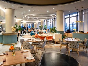 Ein modernes Restaurant mit runden Tischen, blauen Stühlen und großen Fenstern.
