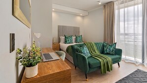 Een moderne slaapkamer met een bed, een groene bank, bureau met laptop, potplant en een groot raam met gordijnen.
